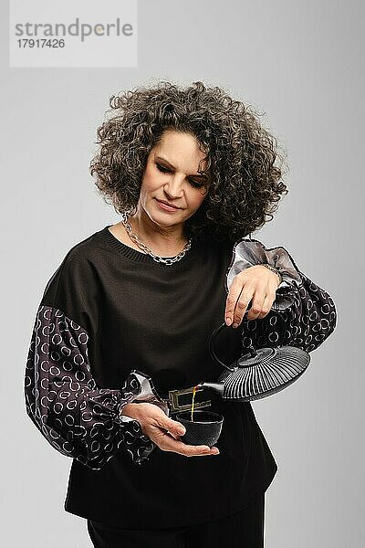 Senior Frau mit grauen Haaren gießt Tee in der Tasse über grauen Studio Hintergrund