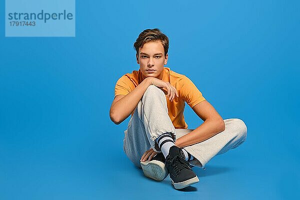 Junger Mann in orangefarbenem T-Shirt und weißer Jogginghose sitzt auf dem Studioboden und stützt sein Kinn vor blauem Hintergrund auf