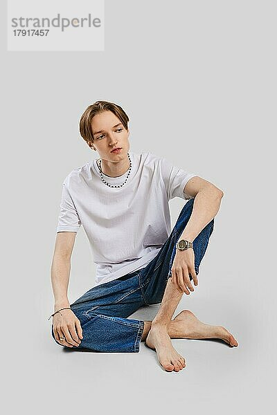 Junger barfüßiger Mann sitzt in T-Shirt und Jeans auf dem Boden im Studio