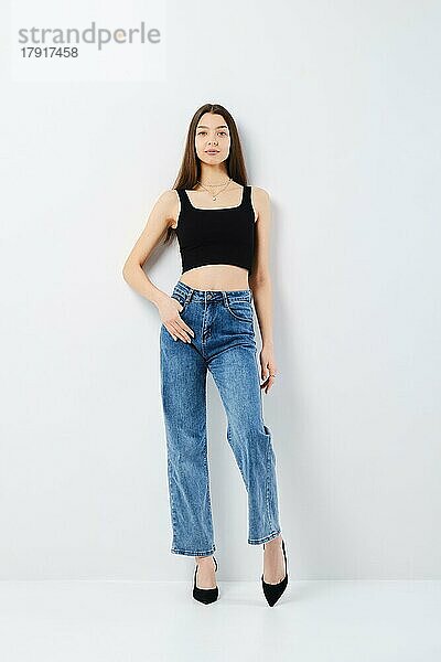 Ganzkörperporträt einer jungen Frau in schwarzem Tanktop und weiten Jeans  die sich an die weiße Studiowand lehnt