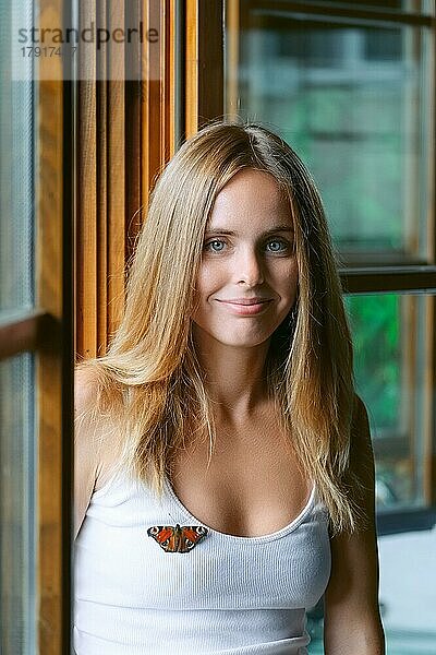 Glückliches lächelndes Mädchen sitzt in der Nähe des Fensters und Schmetterling sitzt auf ihrem T-Shirt