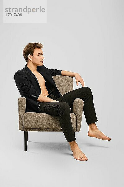 Glücklicher junger Mann in schwarzem Anzug  der in einem Sessel im Studio sitzt