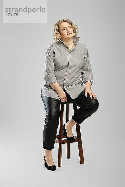 Ältere Frau in grauem Hemd und Kunstlederhose sitzt auf einem hohen Holzstuhl vor grauem Studiohintergrund