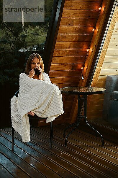 Hübsche junge Frau sitzt in einem Sessel auf der Terrasse eines Landhauses  eingewickelt in eine Decke  mit einer Tasse beruhigenden Tees und genießt die frische Luft