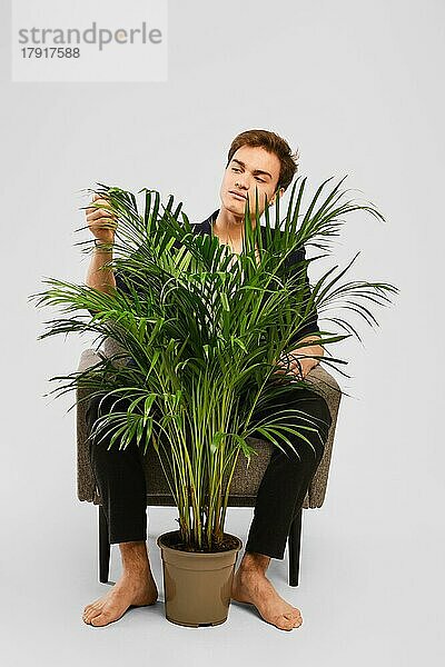 Barfüßiger junger Mann in schwarzem Anzug  der in einem Sessel sitzt und eine Zimmerpflanze im Studio betrachtet