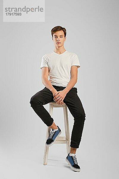Junger gutaussehender Mann in schwarzer Hose und weißem Hemd sitzt auf einem hohen Holzstuhl im Studio