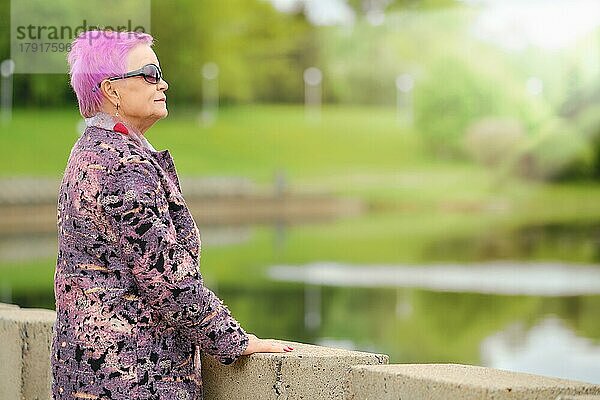 Lustige ältere Frau mit kurzen rosa Haaren und Wollmantel am Flussufer im Stadtpark stehend