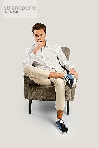 Nachdenklicher junger  gutaussehender Mann in beiger Hose und weißem Hemd sitzt in einem Sessel im Studio