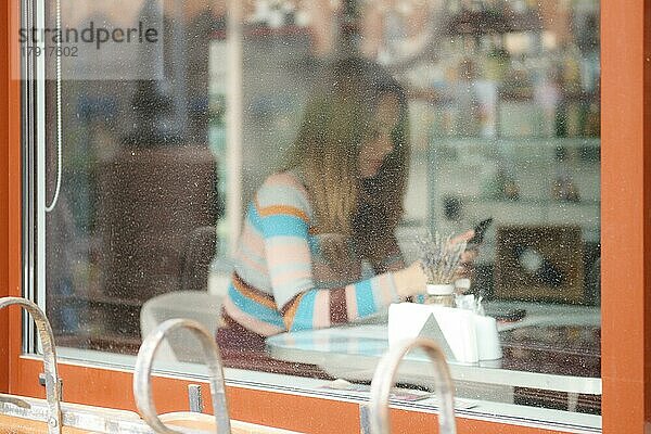 Unscharfes Foto durch das Fenster eines Mädchens in einem Café mit Mobiltelefon