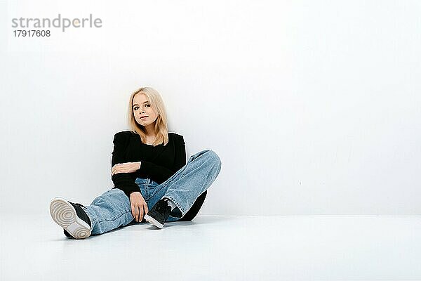 Nettes Mädchen in Tank Top  Jacke und Jeans posiert auf dem Boden sitzend in einem weißen Studio