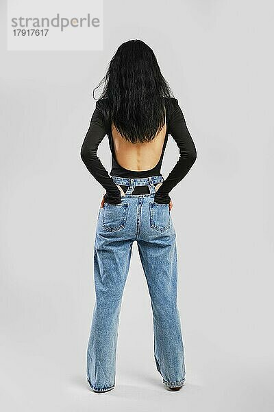 Rückenansicht einer barfuß gehenden Frau mit Händen in den Taschen im Studio in einem schwarzen Body mit langen Ärmeln und einer übergroßen Jeans auf grauem Hintergrund