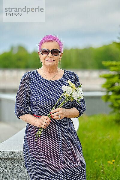 Porträt einer lächelnden reifen Frau mit kurzen rosa Haaren  die in der Nähe eines Tannenbaums auf der Straße steht