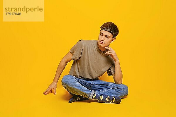 Fröhlicher junger Mann in Alltagskleidung  der in einem hellgelben Studio auf dem Boden sitzt