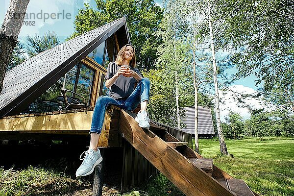 Glückliche Frau sitzt mit einer Tasse Tee auf der Terrasse eines Blockhauses und genießt die frische Luft und das warme Wetter