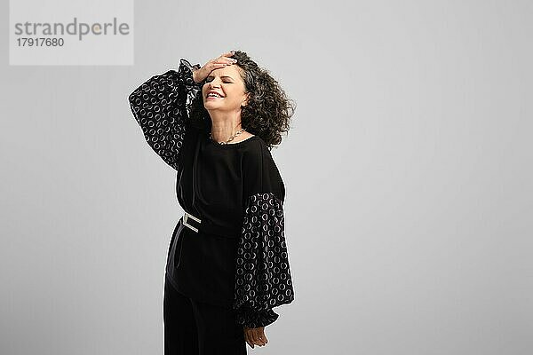 Sehr glückliche ältere Frau mit üppigen lockigen Haaren lachend im Studio vor grauem Hintergrund