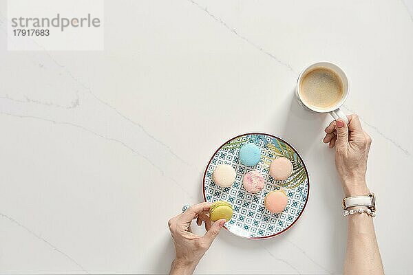Draufsicht auf bunten Kuchen Makronen auf einem Teller und weibliche Hand mit Tasse Kaffee