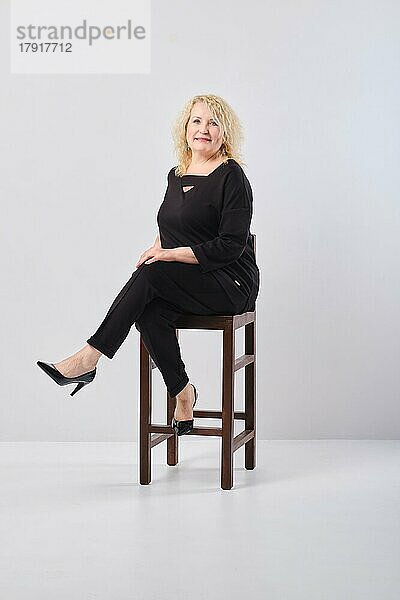 Frau mittleren Alters in schwarzem Outfit sitzt auf einem Stuhl in einem weißen Studio