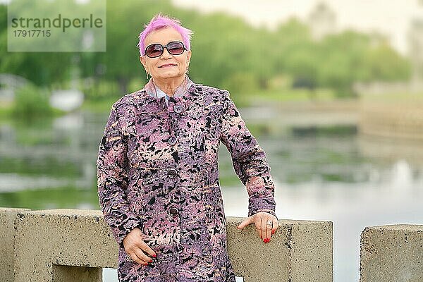 Aktive ältere Frau mit fröhlichem Lächeln steht am Flussufer im Stadtpark