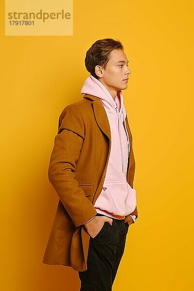 gutaussehender  stilvoller Mann in braunem Trenchcoat und rosa Sweatshirt mit Kapuze im Profil vor gelbem Studiohintergrund stehend