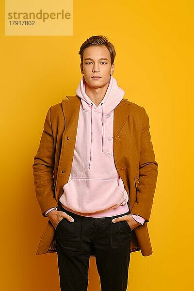 gutaussehender stilvoller Mann in braunem Mantel und rosa Kapuzenpulli  der mit den Händen in den Taschen vor einem gelben Studiohintergrund posiert