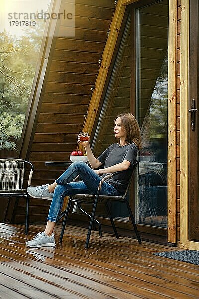Verträumte Frau sitzt im Korbstuhl mit einem Glas Bourbon auf der Holzterrasse nach dem Regen und genießt die Herbstsonne