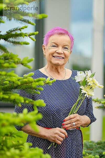 Porträt einer lächelnden reifen Frau mit kurzen rosafarbenen Haaren  die in der Nähe eines Tannenbaums mit Frühlingsblumen in den Händen im Freien steht