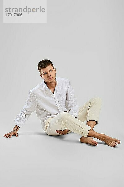 Ganzes Studio-Porträt eines jungen barfüßigen Mannes in weißem Hemd und elfenbeinfarbenen Hosen  der auf dem Boden sitzt