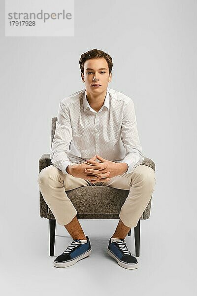 Nachdenklicher junger  gutaussehender Mann in beiger Hose und weißem Hemd sitzt in einem Sessel im Atelier  er hat die Finger verschränkt und schaut geradeaus
