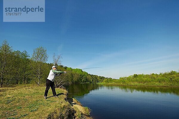 Angler mit einer Spinnrute fangen Fische auf einem Fluss an einem sonnigen Sommertag mit grünen Bäumen im Hintergrund. Wochenendaktivität im Freien. Foto mit geringer Schärfentiefe bei weit geöffneter Blende aufgenommen