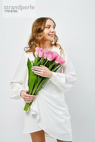 Lächelnde junge Frau in weißem Kleid posiert mit Tulpenstrauß in der Hand vor weißem Hintergrund und schaut zur Seite