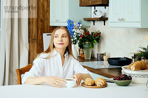 Positive junge Frau sitzt hinter dem Küchentisch mit einer Tasse Kaffee