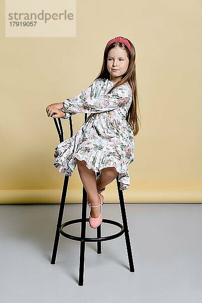 Nettes kleines Mädchen in Sonnenkleid und Haarband sitzt auf einem hohen Stuhl im Studio über blassgelben Hintergrund
