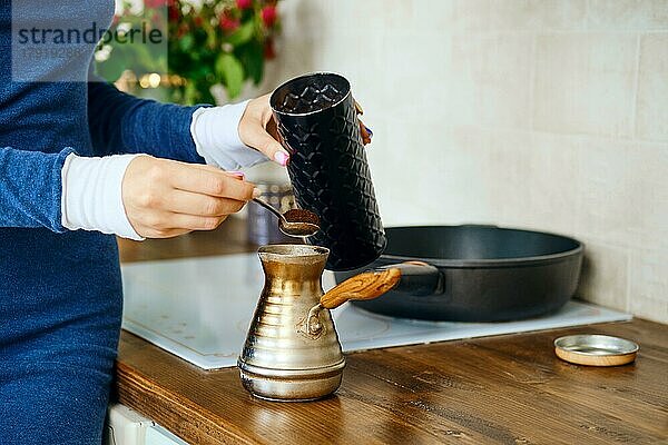 Nahaufnahme einer weiblichen Hand mit Löffel mit gemahlenem Kaffee in der Nähe eines Turks. Kaffeezubereitung in Metallkanne