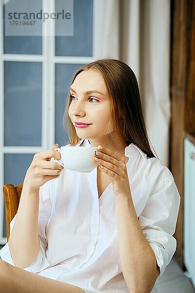 Nachdenkliche junge Frau im weißen Hemd in der Küche hält eine Tasse Kaffee