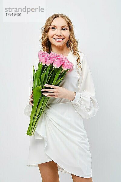 Nette junge fröhliche Frau im weißen Kleid posiert mit Blumenstrauß von Tulpen in der Hand über weißem Hintergrund