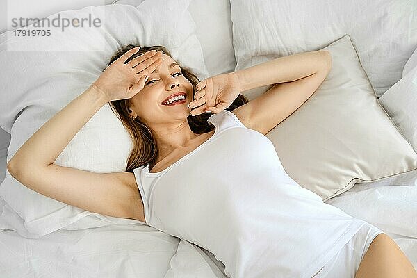Junge positive Frau in weißem Tank Top und Höschen im Bett deckt Gesicht mit Händen und lächelnd