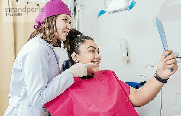 Weibliche Patientin prüft Zähne nach der Zahnbehandlung in der Zahnklinik. Zahnarzt mit zufriedener Patientin lächelnd am Zahnspiegel  Zufriedene Patientin in Zahnklinik schaut in Spiegel
