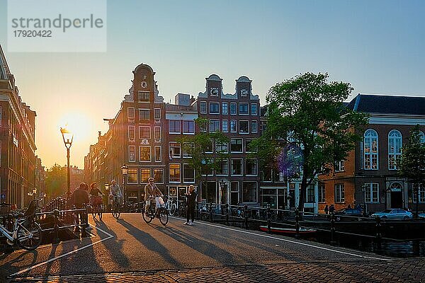 AMSTERDAM  NIEDERLANDE  20. MAI 2018: Menschen auf Fahrrädern auf der Amsterdamer Brücke über die Gracht mit Häusern bei Sonnenuntergang. Amsterdam  Niederlande  Europa