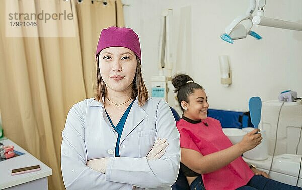 Porträt einer jungen Zahnärztin in einer modernen Zahnklinik. Porträt einer Zahnärztin mit verschränkten Armen vor einem Patienten  Lächelnde Zahnärztin in einer Klinik  Nahaufnahme einer Zahnärztin mit verschränkten Armen