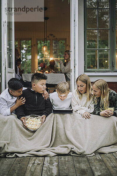 Aufgeregte Freunde mit Decke und Popcorn  die auf der Veranda sitzend Tablet-PC schauen