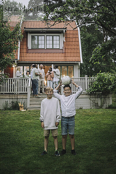 Playful Jungen mit Fußball stehen in Hinterhof mit Familie auf Veranda im Hintergrund