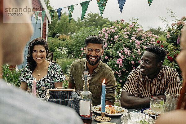 Mann lacht  während er mit männlichen und weiblichen Freunden auf einer Party am Tisch sitzt