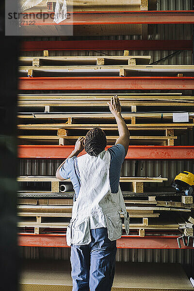 Rückansicht eines männlichen Zimmermanns  der ein Holzbrett auf einem Palettenstapel in einem Lagerhaus sucht