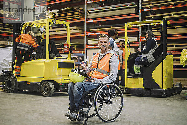 Ganzes Porträt eines lächelnden Zimmermanns mit digitalem Tablet  der auf einem Rollstuhl in einem Lagerhaus sitzt