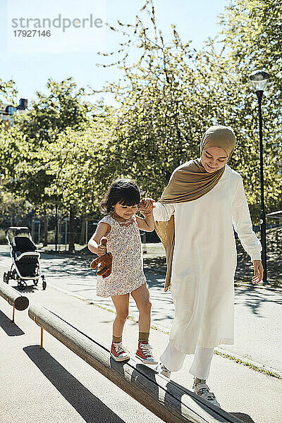 Mutter hält die Hand ihrer Tochter und balanciert auf einer Bank im Park an einem sonnigen Tag