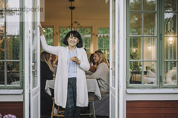 Porträt einer Frau mit Strickjacke  die ein Glas in der Hand hält und an der Tür steht