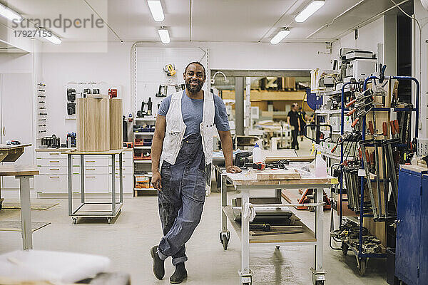 Porträt eines lächelnden Tischlers in voller Länge  der auf einem Bein steht und sich in der Werkstatt auf eine Werkbank stützt