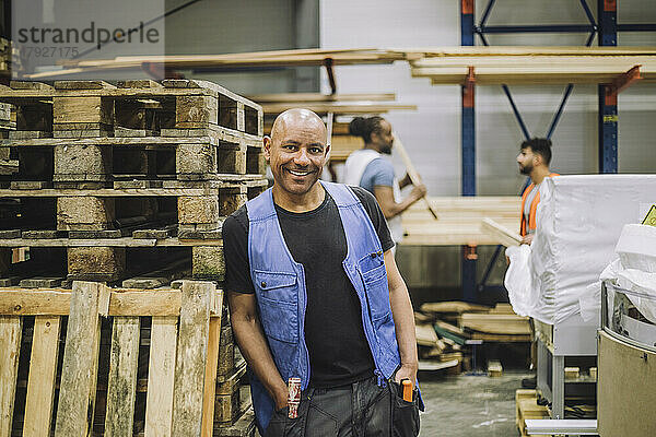 Lächelnder glatzköpfiger Tischler mit Händen in den Taschen  der sich auf ein Holzregal in einem Lagerhaus stützt