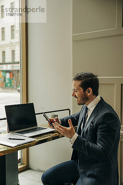 Lächelnder Geschäftsmann  der ein Mobiltelefon hält  während er über drahtlose In-Ear-Kopfhörer im Büro spricht
