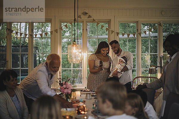 Glückliche Mutter zeigt ihrer Tochter  die vom Vater gehalten wird  das Smartphone während einer Dinnerparty zu Hause